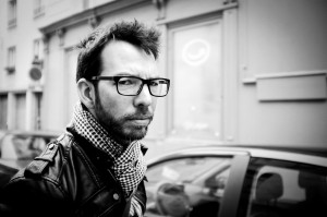 A man with glasses, a beard, and a checkered scarf looks seriously at the camera on a city street. Captured in black and white, this portrait embodies the style of a photographe de rue, with blurred cars and buildings behind him.
