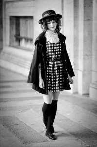 A woman in a robe à carreaux, black coat, knee-high socks, and a hat walks along a city sidewalk. The black and white image shows her looking down with a relaxed expression.