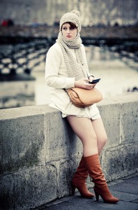 A woman in a cream coat, knit hat, and scarf sits on a stone ledge in the heart of Fashion City, holding a phone. She wears tan knee-high boots and carries a light brown bag. A blurred bridge and water form the backdrop.