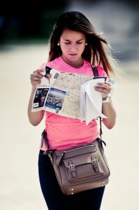A woman wearing a rose pink shirt and carrying a brown purse looks at a carte and some papers outdoors, appearing focused and thoughtful.