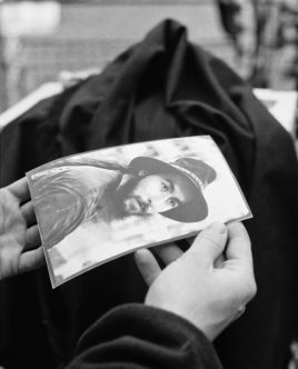 A pair of hands holds a black and white photo of Hans Zeeldieb, wearing a hat and looking directly at the camera. The background is blurred, making him the clear focal point of both the original photo and the photo in hand.