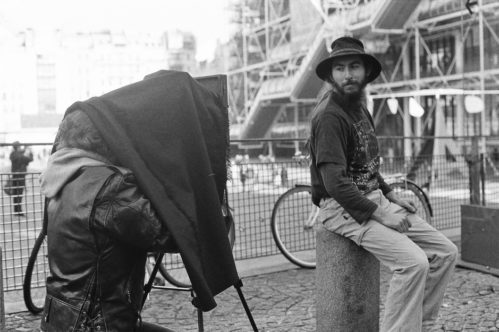 A black-and-white photo shows Hans Zeeldieb sitting on a short pillar outdoors while David Rothan operates a large, old-fashioned camera with a cloth over his head. Bicycles and scaffolding are visible in the background.
