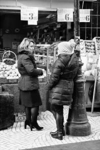 Two women in winter coats share a Causette in front of a fruit and vegetable stall at the marché. One wears heeled boots, the other a beanie and flat boots. Baskets of fresh produce are displayed behind them.