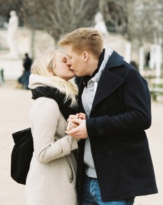 A young couple of jeunes amoureux shares a kiss outdoors in winter clothing. The woman holds a phone and wears a light coat and scarf, while the man wears a dark coat. Blurry trees and statues are in the background.