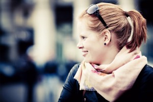 A lovely young woman with red hair tied back, wearing sunglasses on her head, a light scarf, and a black jacket, smiles while looking to the side. The blurred background hints at the charm of Paris, emphasizing her face.