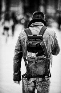 A man in a leather jacket and jeans walks away from the camera, a small dog with les yeux du chat peeking out of his backpack. The black-and-white image captures an urban moment outdoors.