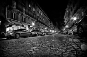 A black and white photo of a deserted cobblestone street in Paris at nuit, lined with parked cars and tall buildings, streetlights casting a glow and a restaurant visible on the left.