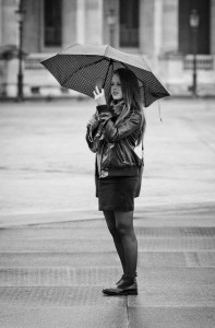 Sous le parapluie, a woman stands outdoors on a wet pavement, holding an open umbrella above her head. She is wearing a leather jacket, skirt, and boots. The image is in black and white, with blurred buildings in the background.