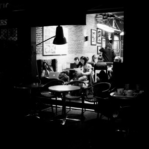 A black-and-white photo of people inside a cozy café. Some are working on laptops, others are chatting like friends after a surf de nuit. Empty tables and chairs sit in the foreground; framed pictures hang on softly lit brick walls.