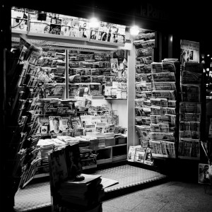 Black-and-white photo of a kiosque de nuit at night, filled with neatly arranged newspapers and magazines on racks and shelves, illuminated by bright lights. The scene appears busy and packed with various publications.