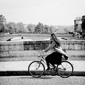 A woman in a robe noire rides a bicycle along a stone bridge, talking on her phone. Trees and buildings line the distant background, and a river runs beneath the bridge. The black and white image captures the quiet vent of city life.