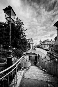 A black and white photo of the steep escalier de la station Passy with a lamppost on the left, leading down to a narrow street or railway station, surrounded by buildings and trees under a dramatic cloudy sky.