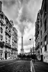 Black and white photo of a cobblestone street in Paris, framed by tall buildings, with the Eiffel Tower centered in the background under a dramatic, cloudy sky.