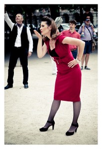 A woman in a vibrant red dress and black heels stands on a path at Place des Vosges, blowing a kiss. Behind her, a man in a white shirt and dark vest raises his hand. Other people are visible in the background, including someone in a purple shirt holding a camera. It feels like the start of an impromptu flash mob.