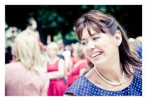 A woman with brown hair in a ponytail smiles while looking to her right. She is wearing a navy blue dress with white polka dots and a pearl necklace. The background is blurred, showing people in red and pink clothing, suggesting an outdoor social event, possibly a flash mob at Place des Vosges.