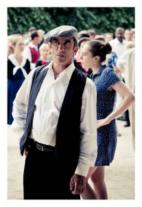 A man wearing a flat cap, white shirt, and dark vest stands in the foreground with a serious expression. Behind him, at Place des Vosges, a woman in a blue polka-dot dress and others in various attire are engaged in conversation or other activities. The background hints at an impromptu flash mob amidst the leafy outdoor setting.
