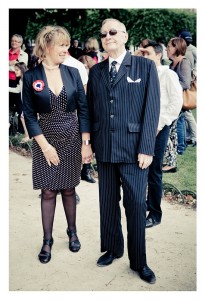 A man in a pinstripe suit and sunglasses stands holding hands with a woman in a black polka dot dress and jacket at Place des Vosges. They are outdoors with a crowd of people, possibly part of a flash mob. The woman is looking at the man and smiling.