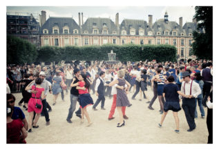 People are dancing in pairs in an outdoor square with a fountain in the center, resembling a vibrant flash mob. The crowd is diverse, with individuals dressed in vintage attire. Historic buildings serve as the backdrop at Place des Vosges, and the atmosphere appears festive and lively.