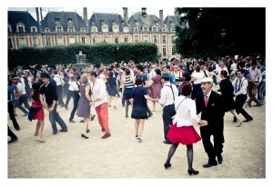 A large group of people are dancing together outdoors in front of a historic building at Place des Vosges, with a fountain and trees in the background. The dancers, part of a flash mob, are dressed in various attire, and there is a lively atmosphere. Some onlookers are visible in the background, observing the scene.