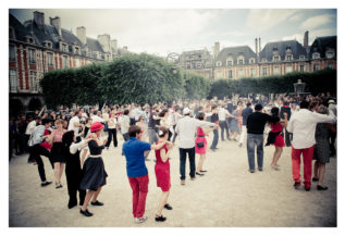 A large group of people dancing in an open square surrounded by historic buildings, likely Place des Vosges. Many dancers hold hands or dance in pairs. The scene feels like a flash mob, festive with participants wearing colorful clothing. An audience watches from the sides, green trees in the background.