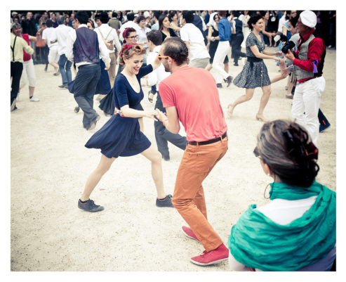 A large group of people dancing energetically outdoors at Place des Vosges. In the foreground, a man in an orange shirt and pants dances with a woman in a navy blue dress. Other dancers surround them, dressed in various casual and vintage-style outfits. The flash mob atmosphere is lively and joyous.