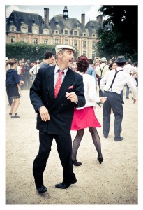 A man in a black suit and white hat dances outdoors with a flash mob dressed in vintage attire at Place des Vosges. People in the background, including a woman in a red skirt, join the lively dance. The scene buzzes with energy, with historic buildings and trees framing the backdrop perfectly.