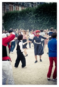 A lively outdoor scene shows a flash mob dancing exuberantly. A woman in a black dress and red hat stands out at the center, surrounded by dancers dressed in various casual attire. Trees and a building with red brick and white windows are in the background, reminiscent of Place des Vosges.