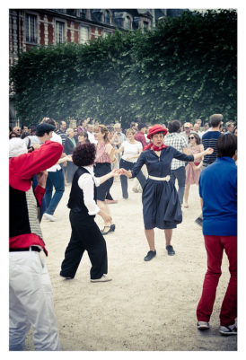 A lively outdoor scene shows a flash mob dancing exuberantly. A woman in a black dress and red hat stands out at the center, surrounded by dancers dressed in various casual attire. Trees and a building with red brick and white windows are in the background, reminiscent of Place des Vosges.