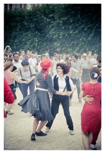 A group of people dancing outdoors at Place des Vosges. The retro-themed scene features vintage clothing; one person in a gray dress and red hat dances with another in a white shirt and black suspenders. It's a lively flash mob, with a crowd gathered in the background, some smiling and others taking photos.