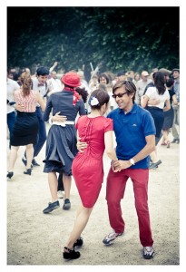 A group of people are outdoors, dancing energetically in what appears to be a spontaneous flash mob. The couple in the foreground is dressed casually; the woman in a red dress and the man in a blue polo shirt with red pants. Other dancers at Place des Vosges are visible in the background, enjoying the lively atmosphere.