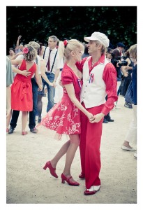 A couple dressed in matching red-themed outfits, including red shoes and a white vest for the man, dance together outdoors among other dancing couples at Place des Vosges. The woman wears a red dress with white floral patterns. The setting appears lively with a celebratory flash mob atmosphere.