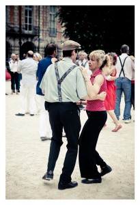 A man and woman dance together outdoors at Place des Vosges. The man wears a cap, suspenders, a light green shirt, and dark pants. The woman wears a red sleeveless blouse and black pants. Other couples are dancing in the background, making it feel like an impromptu flash mob in a park-like setting.
