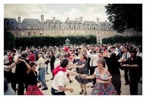 A large flash mob of people, dressed in vintage attire, dance together in the outdoor setting of Place des Vosges with historic buildings in the background. The dancers are wearing colorful polka-dot skirts, hats, and scarves, creating a festive, retro atmosphere.