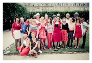 A group of people, some dressed in vintage-style clothing, pose for a photo in an outdoor setting. Many of them are wearing red, white, and blue with accessories like ribbons and hats. The backdrop includes trees, ornate buildings at Place des Vosges, and a grassy area.