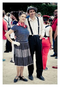 A woman and a man stand side by side, both dressed in vintage clothing, at what appears to be a flash mob. The woman wears a striped top with a polka dot skirt and a red neckerchief, while the man sports a white shirt, black pinstriped pants with suspenders, and a gray hat. They are outdoors at Place des Vosges with a group of people in