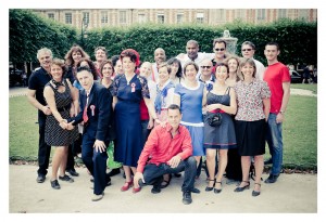 A group of 21 people, dressed in a mix of casual and semi-formal attire, pose together at Place des Vosges with greenery and a fountain in the background. Smiling, they appear to be enjoying a social gathering that looks like it could turn into an impromptu flash mob.