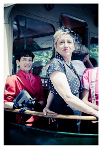 Two women dressed in vintage clothing on a bus, possibly en route to a flash mob. One wears a red jacket with a tie and black gloves, holding a clutch. The other dons a black polka dot dress and headpiece, smiling. A man in vintage attire is partially visible behind them, suggesting Place des Vosges flair.