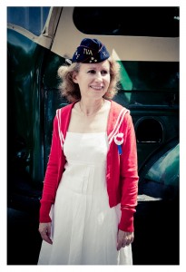 A person with shoulder-length curly hair stands in front of a vintage vehicle at Place des Vosges. They wear a navy blue hat with insignia, a white dress, and a red cardigan with white trim. A blue and red ribbon adorns their outfit, making them look like they just stepped out from a flash mob performance.