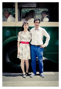 A man and a woman stand next to each other in front of a vintage green bus at Place des Vosges. The woman wears a white dress with a red belt, and the man sports a white shirt with blue pants and a bow tie. Both are smiling with their arms around each other, as if caught in the joy of an impromptu flash mob.