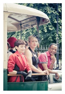 Three people dressed in vintage clothing are standing at the open window of a vintage bus. The person on the left is in a red jacket, the person in the middle is in black, and the person on the right is in a bright pink shirt. They appear to be part of an impromptu flash mob with trees and possibly Place des Vosges in the background.