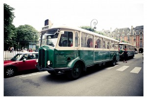 A green and white vintage bus is parked on a street near some historic buildings with ornate facades and chimneys, reminiscent of Place des Vosges. A red car is parked to the left of the bus. Some people, possibly part of a flash mob, are visible around the bus. Trees frame the scene on the left.