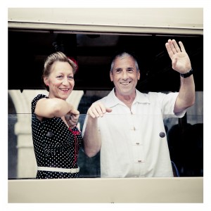 A smiling woman in a black polka dot dress with a red ribbon in her hair and a smiling man in a white shirt are waving from the window of a vehicle. The window is partially open, and they stand close together, perhaps just returning from an impromptu flash mob at Place des Vosges.