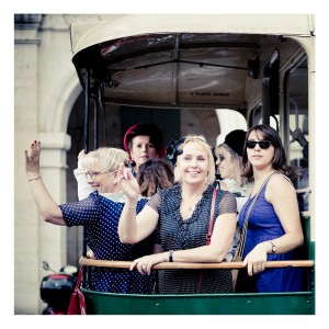 A group of women, dressed in vintage-style clothing, stand and wave from the inside of a charming, open-sided trolley at Place des Vosges. The background shows a building with archways. One woman wears sunglasses, and others have hats and accessories, reflecting a retro fashion flash mob.