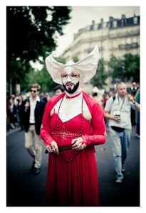 A person dressed in a vibrant red gown and a white headpiece stands in the foreground at an outdoor Gay Pride event. They wear glasses, a necklace of large beads, and have a white and black painted face. Several people and trees are visible in the background.