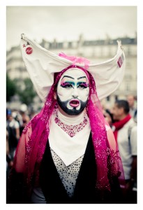 A person dressed in an elaborate costume featuring a white headpiece with pins, bright pink accents, and heavy, dramatic makeup. They are wearing a pink lace veil and white outfit adorned with sequins, standing outdoors against a blurred urban background during the 2014 Gay Pride parade.