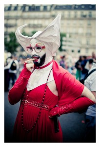 A person dressed in an elaborate costume poses for the camera at a 2014 Gay Pride event. They are wearing a red dress with matching gloves, a beaded necklace, and large glasses. They have face paint and a white headpiece with large, wing-like extensions. The background shows a blurred street scene.