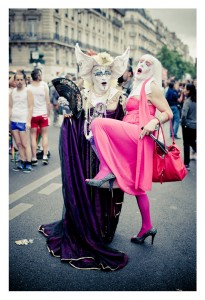 At the 2014 Gay Pride event, two people are dressed in flamboyant costumes. One, in a purple robe and white face paint with bountiful accessories, holds a fan. The other, in a pink dress and matching tights, is being held in a playful pose, leg raised. A lively crowd is in the background.