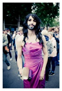 A person with long black hair and a beard is wearing a bright pink strapless dress and holding a metallic clutch. They are standing outside in a crowd of people at the 2014 Gay Pride parade. Trees and other individuals can be seen in the background.