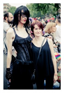 Two individuals pose for a photo at an outdoor event during Gay Pride 2014. The taller person on the left wears a black dress, gloves, and a lace mask. The shorter person on the right wears a black sleeveless top and has short red hair. A crowd is visible in the background.