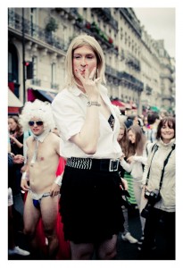 Person dressed in a short black skirt, white blouse, and blonde wig, blowing a kiss towards the camera during the 2014 Gay Pride parade. In the background, several people, including someone dressed in a white wig and minimal clothing, walk along a bustling city street with buildings visible.